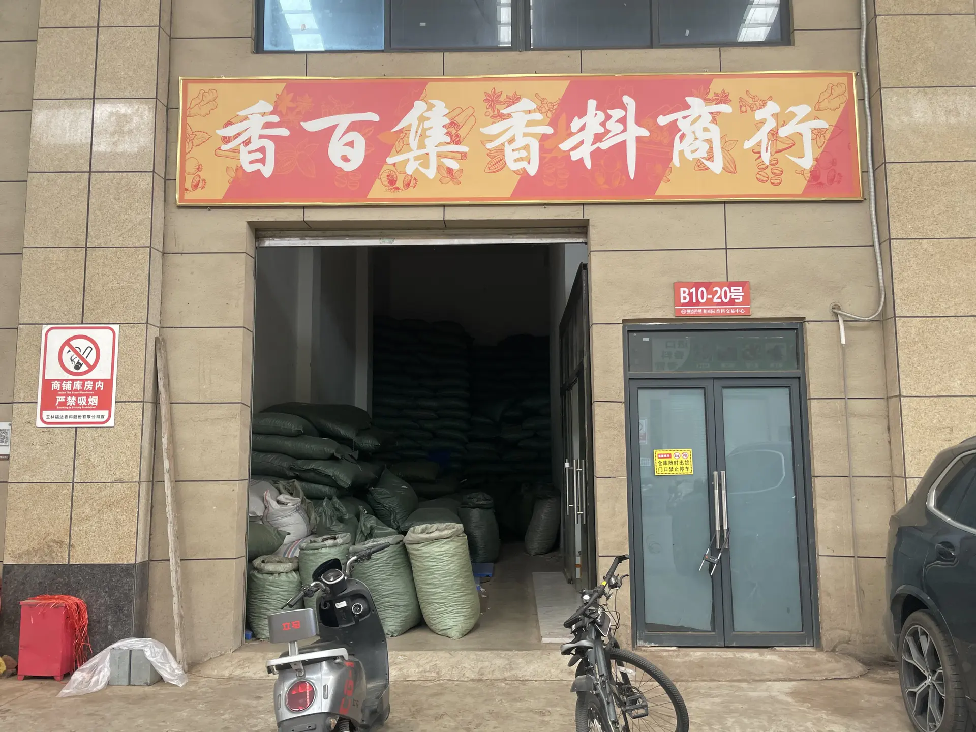 Storefront with the Xiangbaiji spice shop sign above an open roller door, sacks of spice stacked inside, unit number B10-20 beside the door