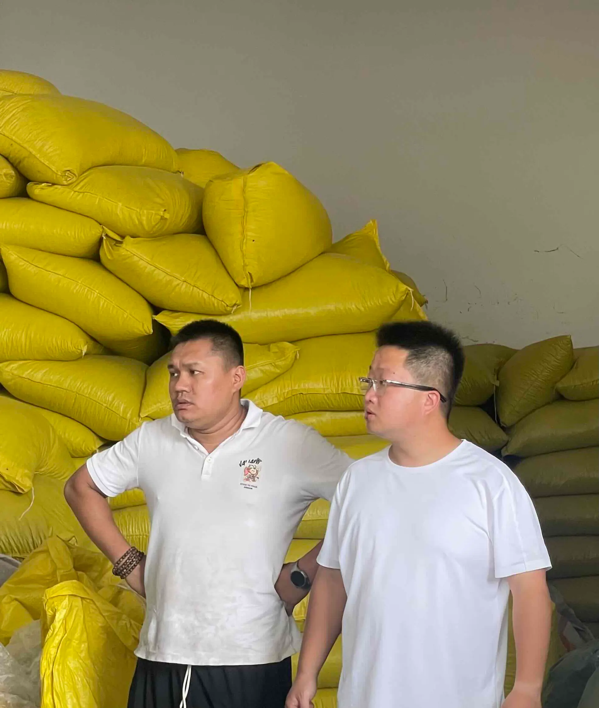 Kun and Chuangui standing in front of stacked yellow sacks of spice, looking at a lot together
